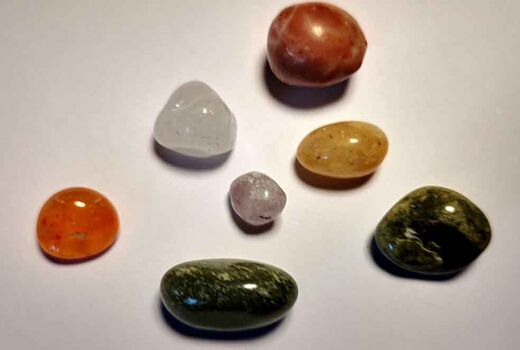 Sieben glatt polierte Trommelsteine liegen mit Abstand zueinander auf einem hellen Untergrund. Zu sehen sind orangefarbene, grüne, gelbe, weiße, transparente und rotbraune Steine in runden und ovalen Formen.