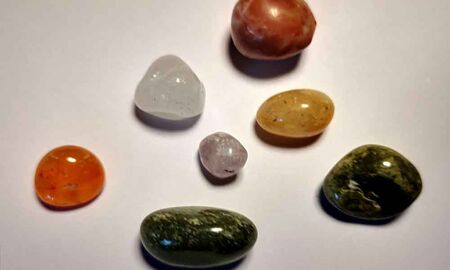 Sieben glatt polierte Trommelsteine liegen mit Abstand zueinander auf einem hellen Untergrund. Zu sehen sind orangefarbene, grüne, gelbe, weiße, transparente und rotbraune Steine in runden und ovalen Formen.